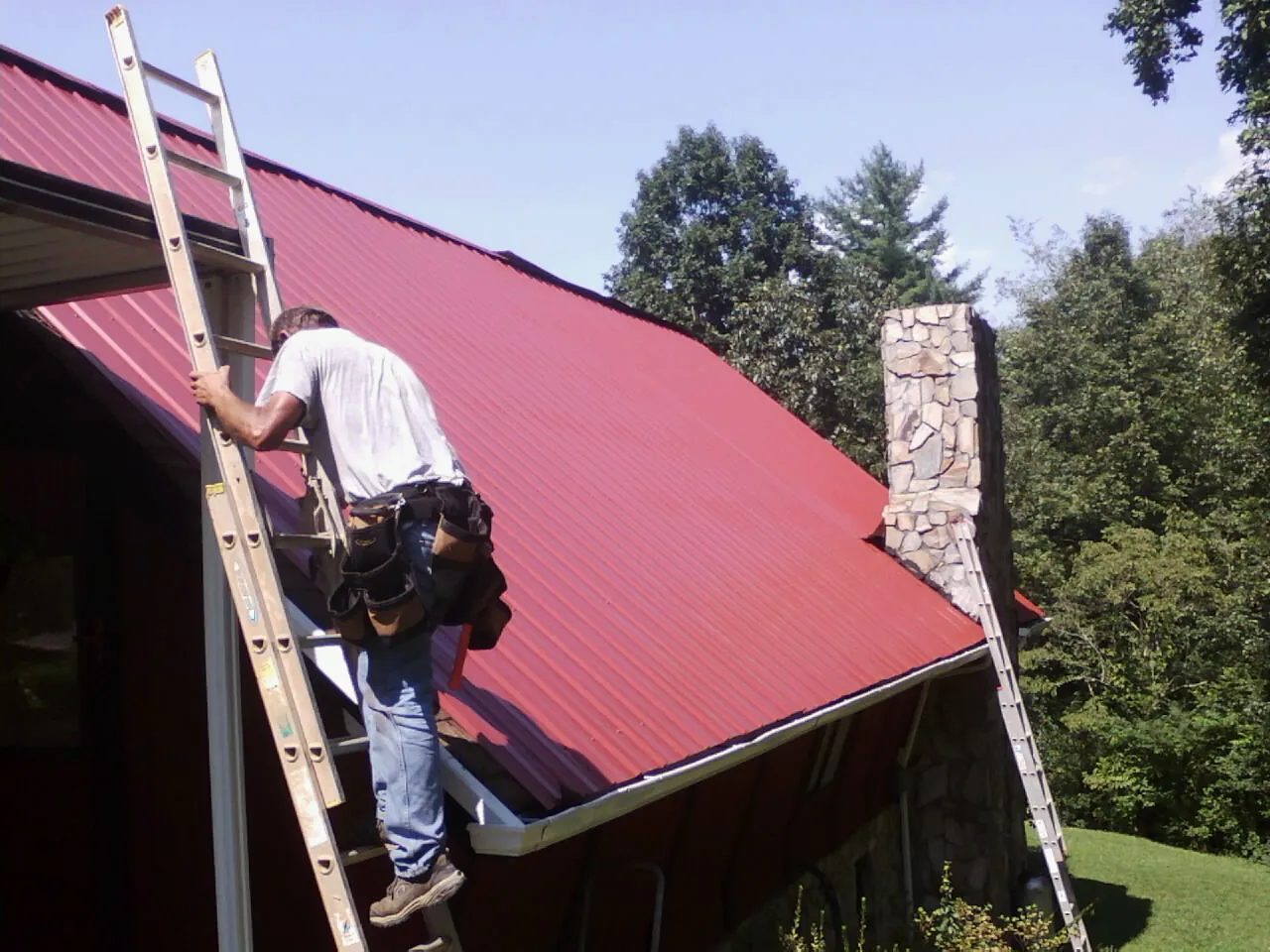 Roofer on a ladder beside a red metal roof and stone chimney — Soffit & Fascia Repair by Roofing Brothers in Kennedyville